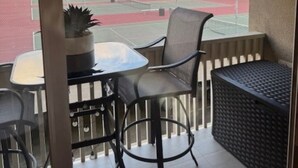 Un balcón con una mesa y sillas, una planta en maceta y una cancha de tenis al fondo.