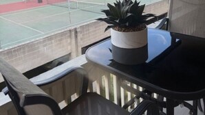 Un balcón con vista a una cancha de tenis, una planta en maceta y una mesa con sillas.
