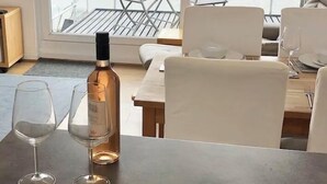 Ein Essbereich mit Tisch für zwei Personen, inklusive Weinflasche, Gläsern, einer Käseplatte mit Trauben und Crackern, und Blick auf Strand und Berge durch große Fenster.