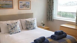 Ein ordentlich bezogenes Bett mit einer karierten Decke und aufgerollten Handtüchern, einer Nachttischlampe und einem Fenster mit Vorhängen.