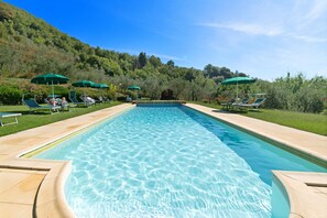 Piscina com água cristalina, rodeada por espreguiçadeiras e guarda-sóis, com um cenário de vegetação exuberante e colinas.
