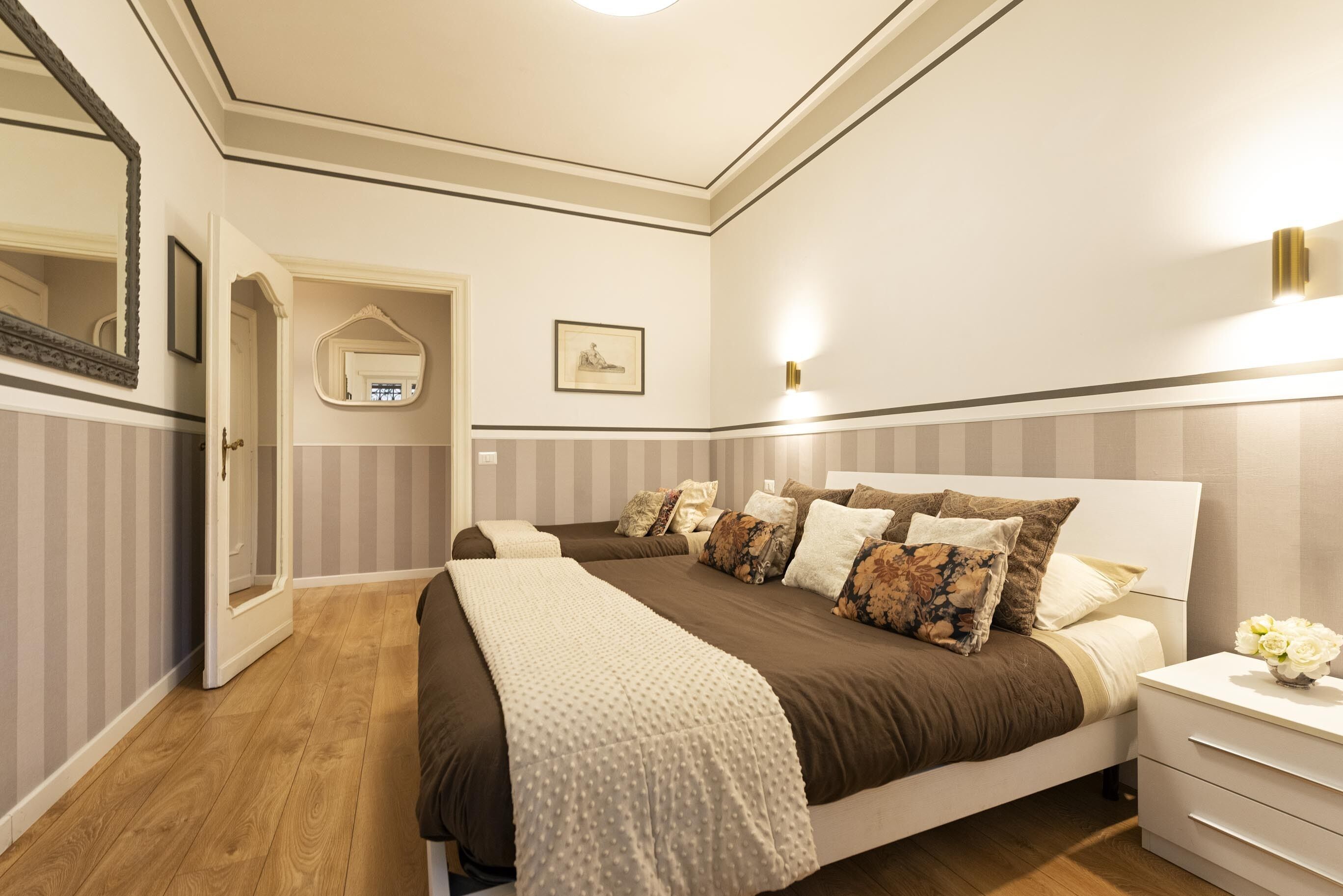 A bedroom with a large bed, a bedside table with a vase of flowers, a mirror, and a framed picture on the wall.