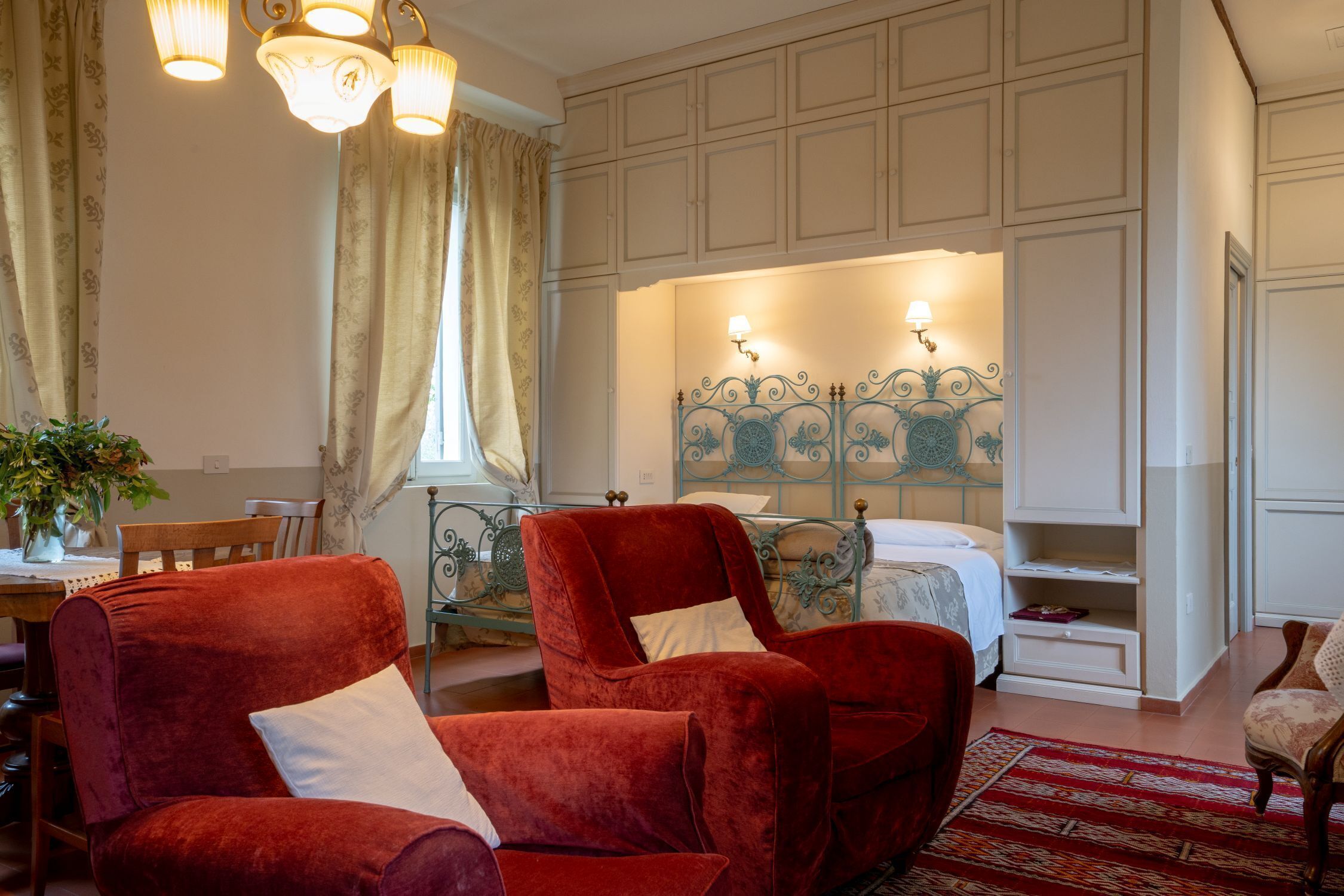 A room with a bed, a red velvet armchair, a dining table with chairs, and a decorative headboard.