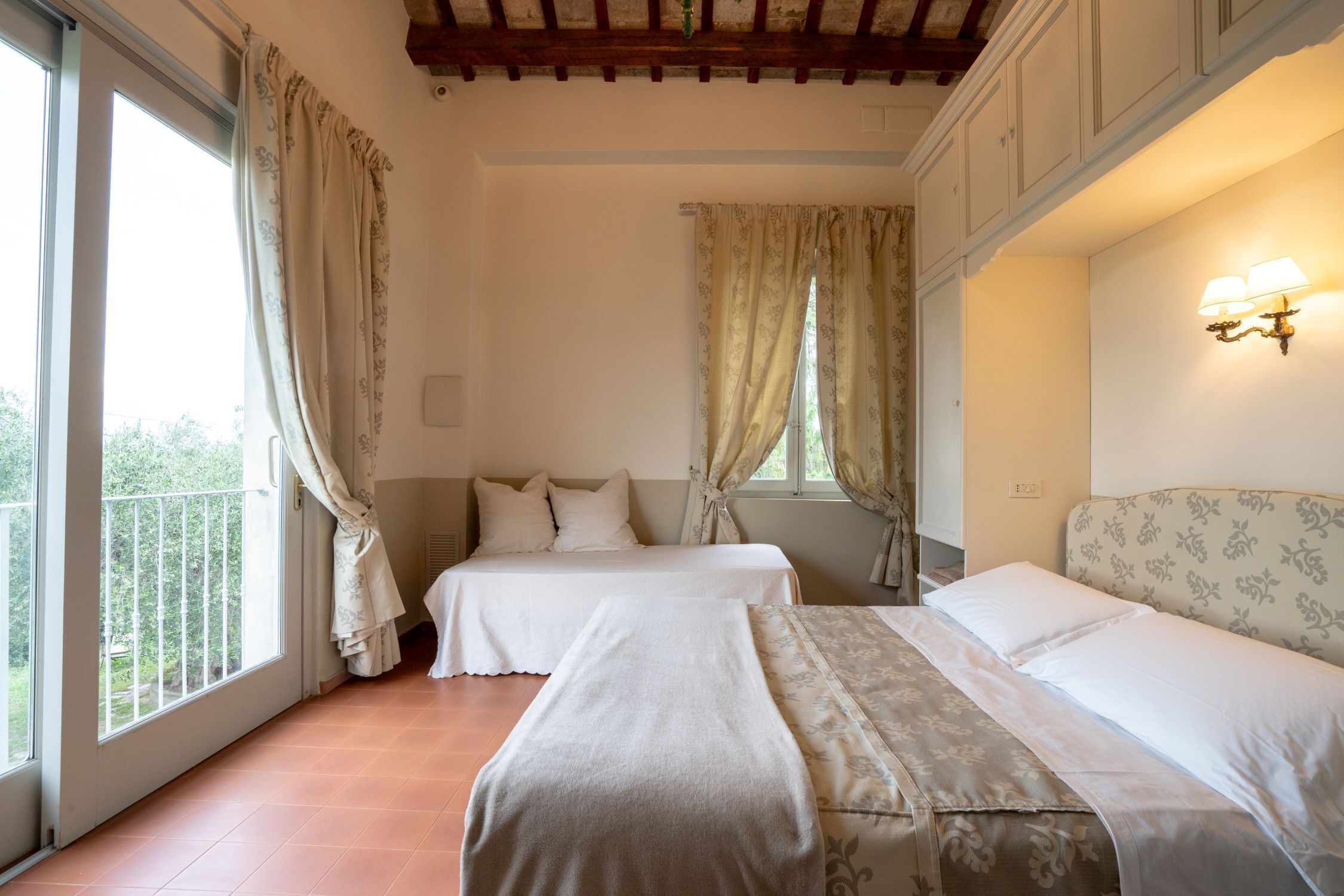 A room with two beds, large windows with curtains, a balcony, and a wall-mounted light fixture.