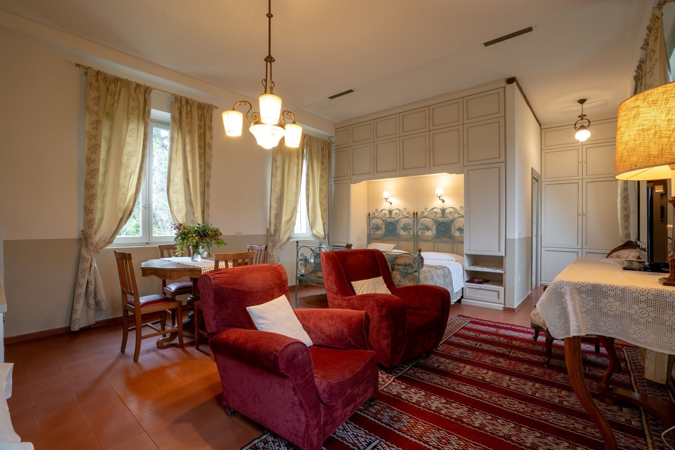 A room with a dining area, a red velvet armchair, a patterned rug, and a bed with a headboard.