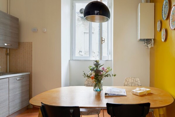 Et spiseområde med et rundt bord, stole, en vase med blomster, et vindue og en vægmonteret lampe.