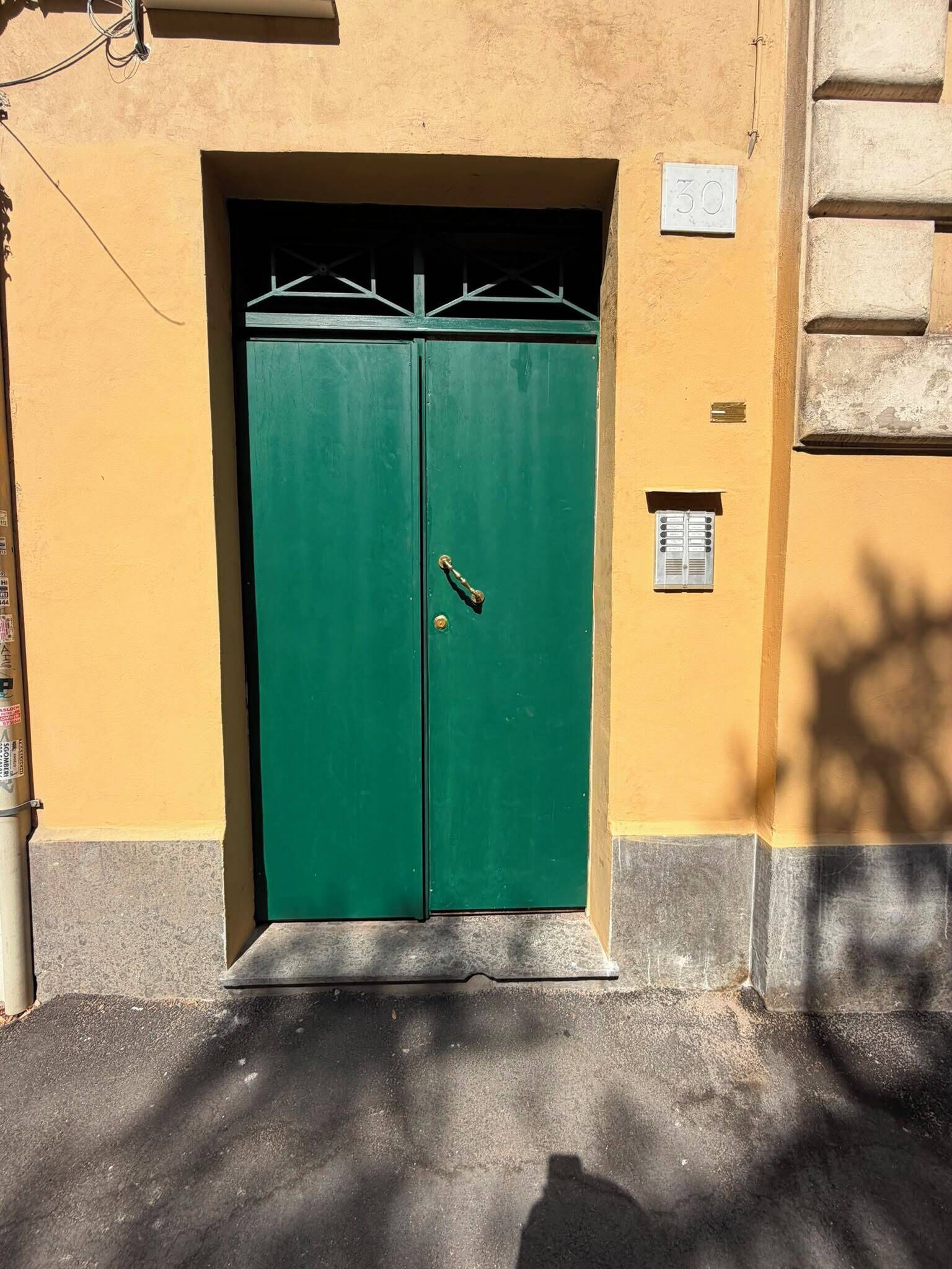 A double green door with a metal handle, set in a yellow building with a small window and a number plate above it.