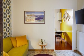 A room with a yellow sofa, a small round table with a plant, a framed picture of a cityscape, and a dining area with a yellow wall and a TV. - TrastevereHome - Peretti Rome (Roma)