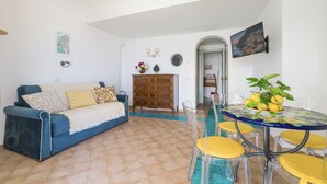 Un soggiorno con un divano blu, una cassettiera in legno, un tavolo in vetro con sedie gialle e vista su una località costiera attraverso un arco.