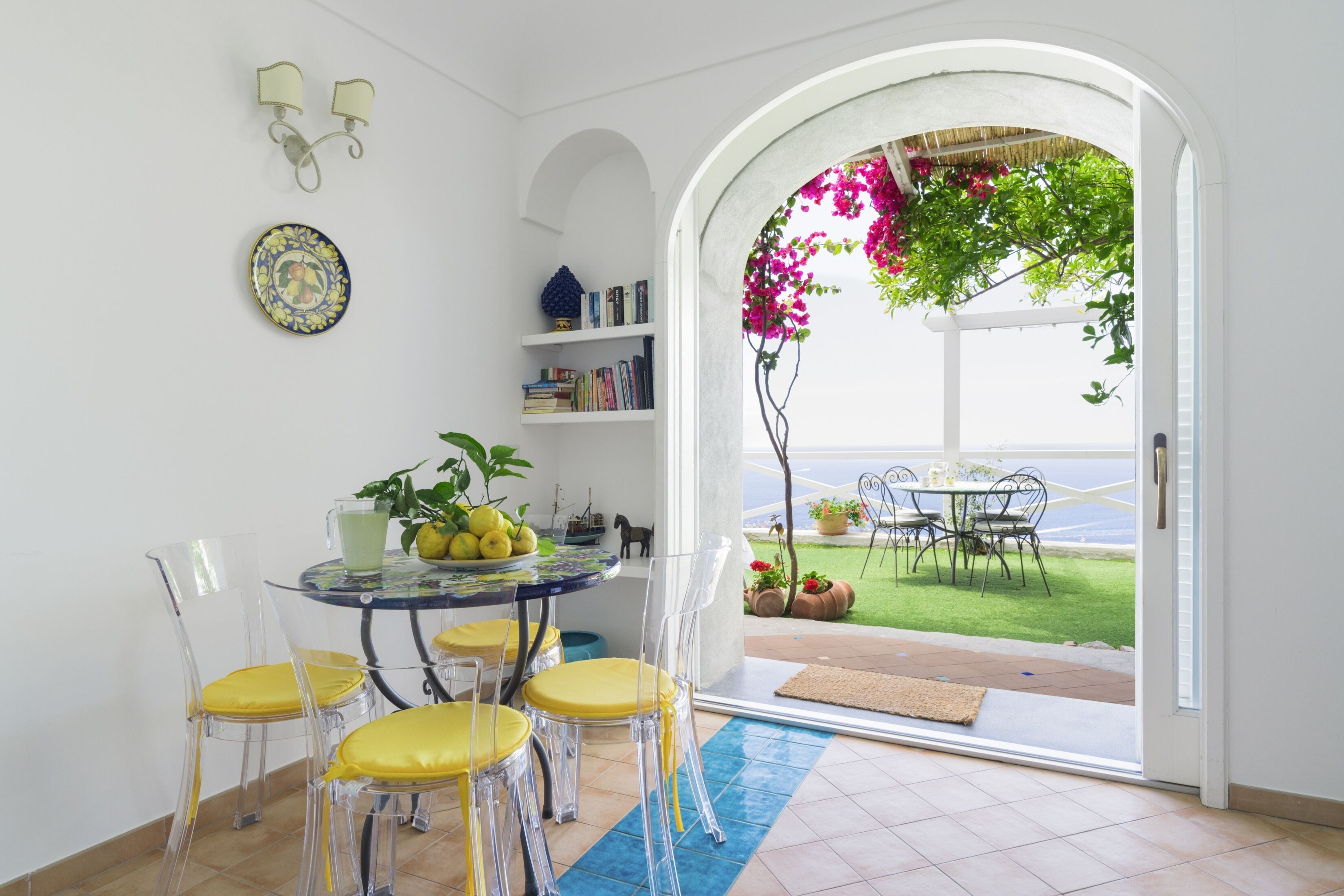 Un comedor con una mesa redonda y sillas, una estantería y vistas al exterior a través de una puerta abierta.