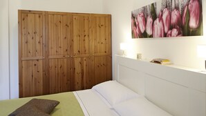Une chambre à coucher avec un lit, des oreillers, une armoire en bois, un tableau de fleurs et une table de chevet avec une lampe et des livres.