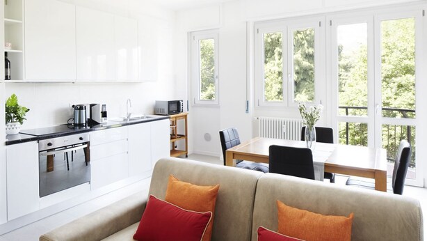 Une cuisine moderne avec des meubles blancs, un coin repas comprenant une table et des chaises, et un canapé agrémenté de coussins rouges.