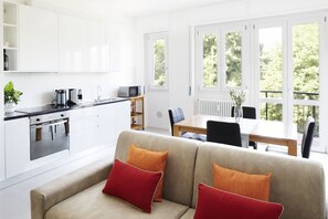 Moderni keittiö, jossa on valkoiset kaapit, ruokailutila pöydän ja tuolien kera sekä punaisilla koristetyynyillä varustettu sohva.