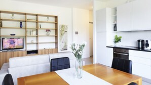 Un salon moderne avec une table à manger en bois, des chaises noires, un canapé blanc et un téléviseur. On y trouve une bibliothèque avec des objets décoratifs et un tableau accroché au mur.