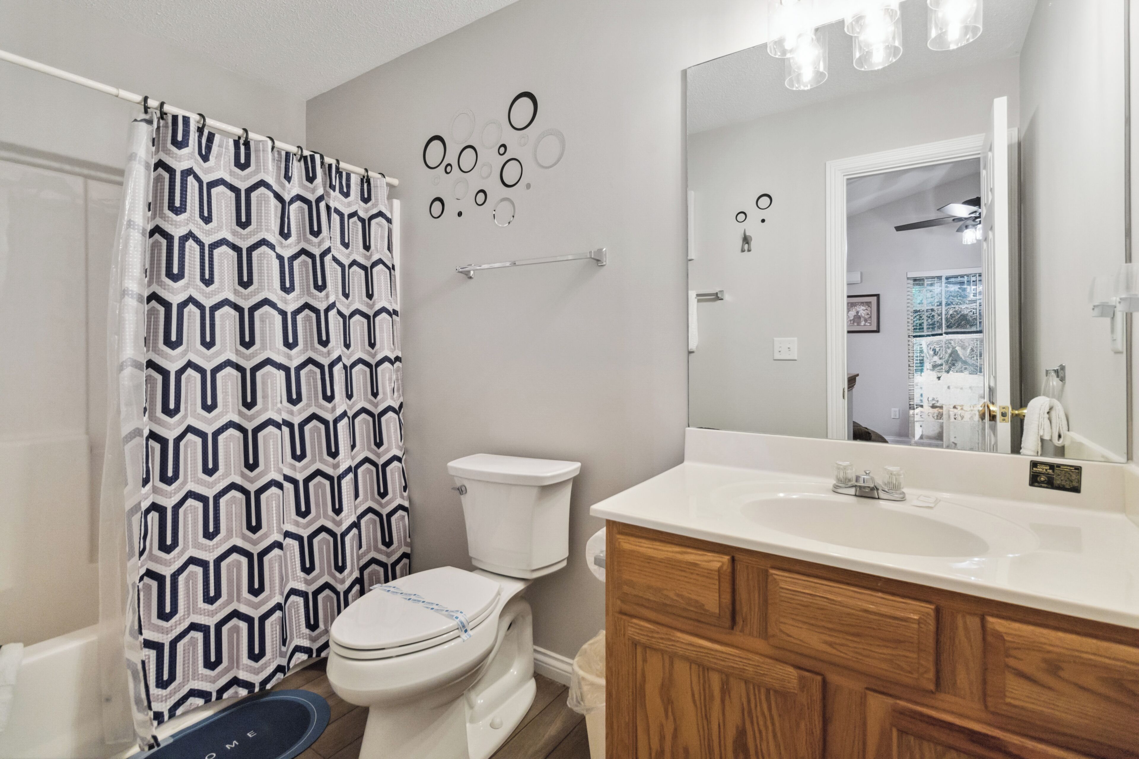 Un baño con inodoro blanco, tocador de madera, lavamanos blanco y cortina de ducha con estampado geométrico.