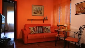 Ein Zimmer mit oranger Wand, einem roten Sofa mit gemusterten Kissen, einem Glastisch mit Stühlen und einem Gemälde über dem Sofa.