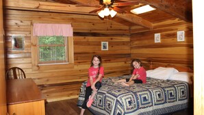 A wooden cabin room with a bed, a ceiling fan, a window with blinds, and a small table. - Wild and Wonderful Wilderness Cabin! (Hico)
