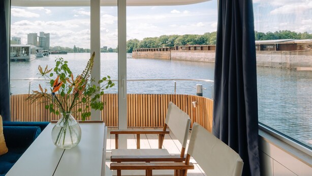 A modern room with a view of a river, a sofa, a table with a vase of flowers, and a wooden railing. - Spreeapartment MARA (Berlin)
