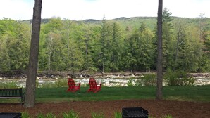 A river with a rocky bed, surrounded by lush green trees and a grassy area with two red Adirondack chairs. - Gorgeous 3BR, 3BA Townhouse Right on the Pemi River - Great Views! (Lincoln)