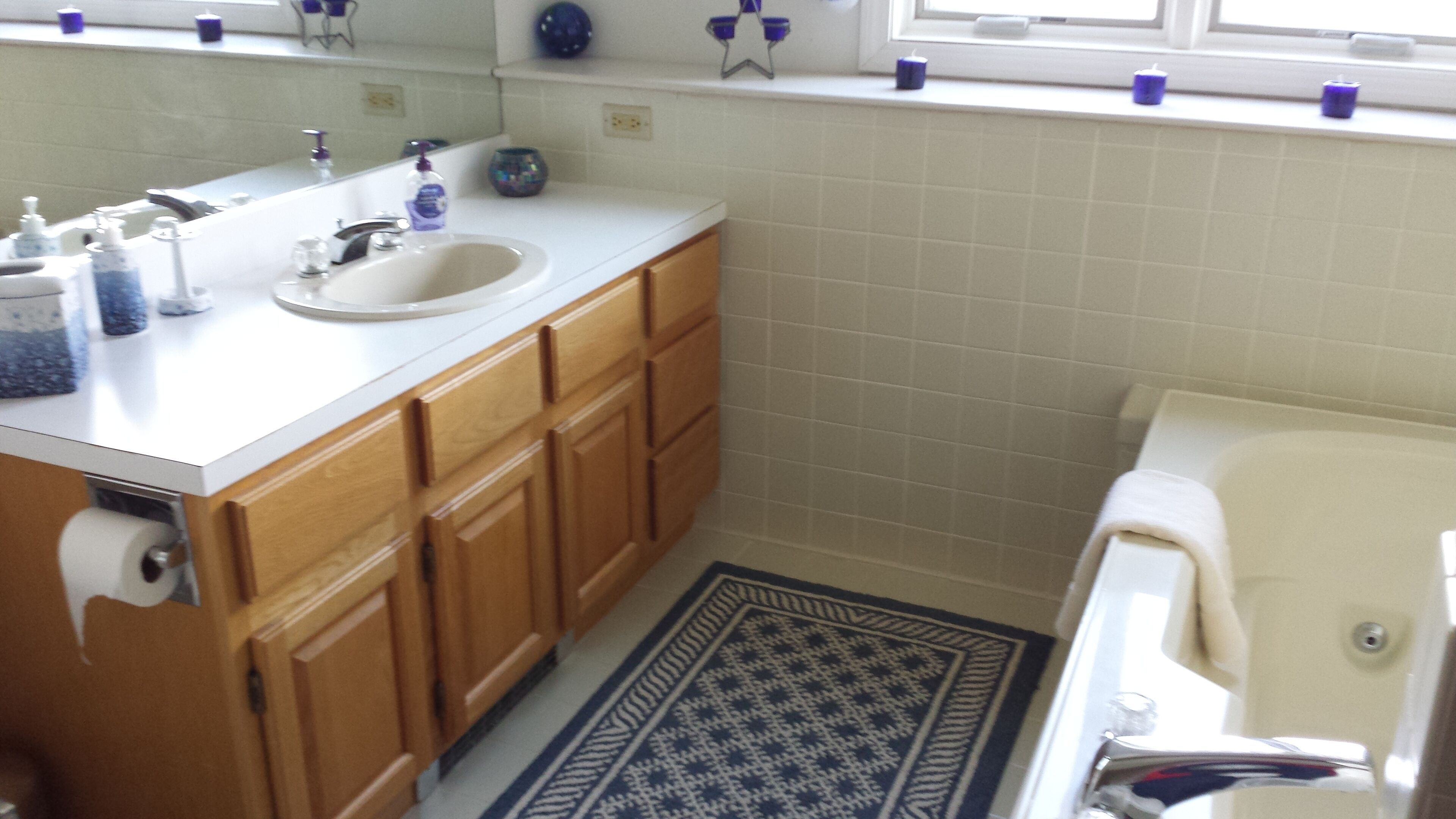 A bathroom with a white sink, wooden cabinets, a patterned rug, and a window with blue candle holders.