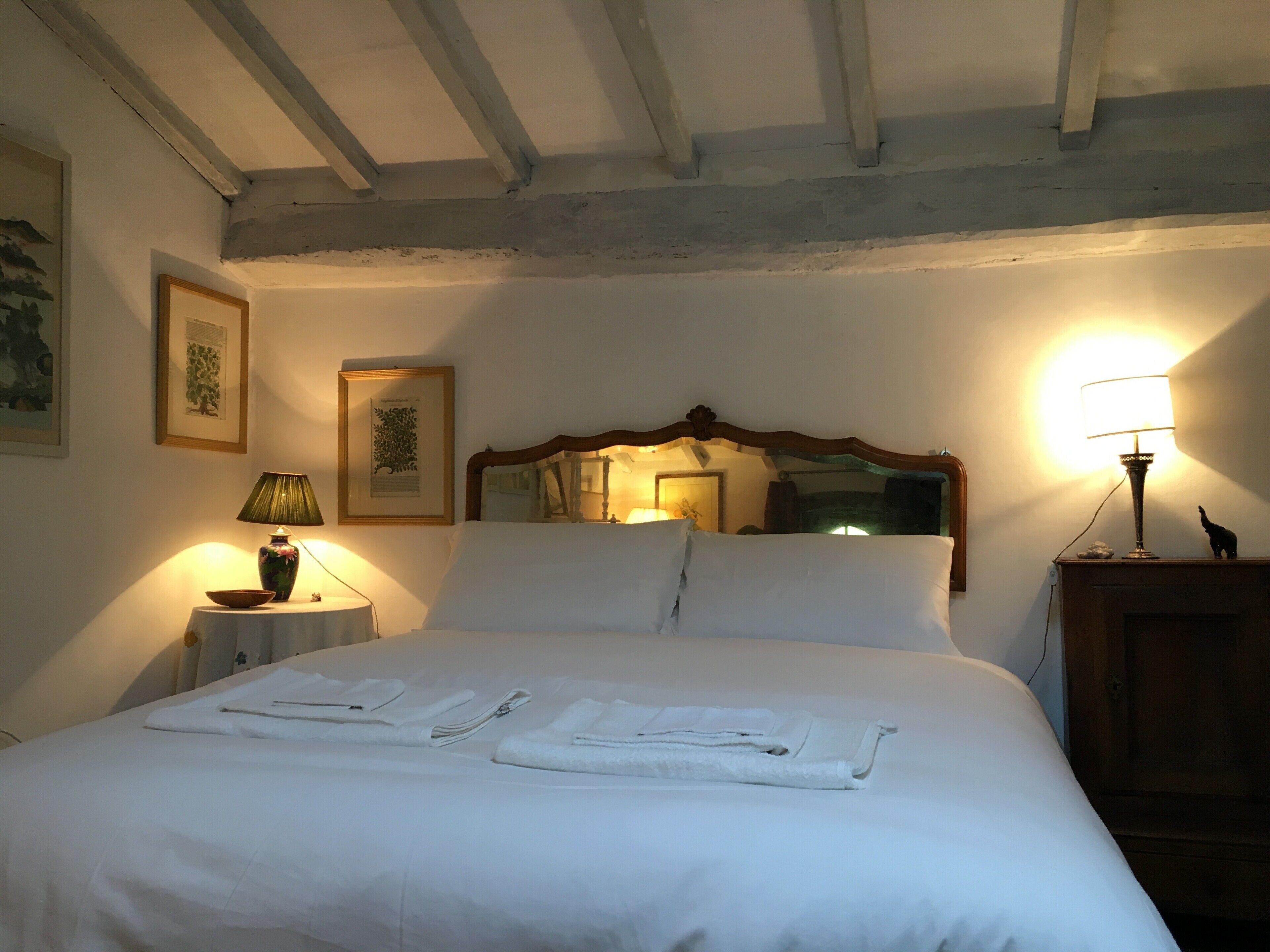 Ein ordentlich bezogenes Bett mit weißen Leinen und Handtüchern, ein hölzernes Kopfende, eine Nachttischlampe und gerahmte Kunstwerke an der Wand.