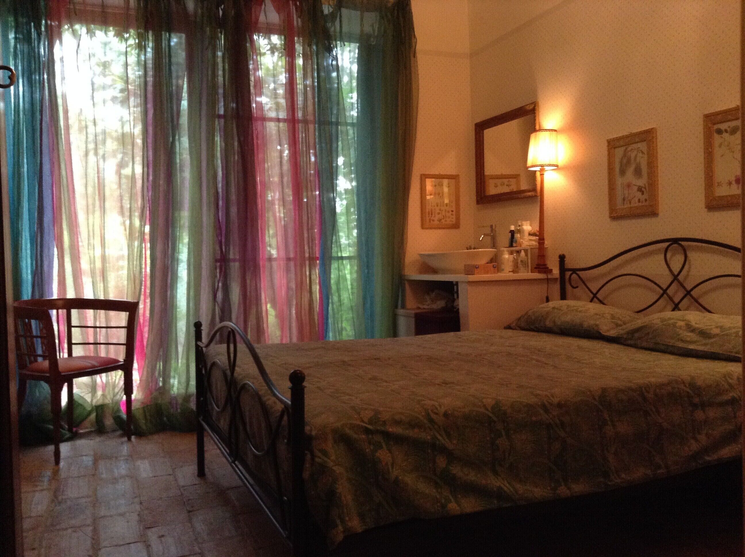 Una camera da letto con un letto, una sedia, un comodino con una lampada, uno specchio e una finestra con tende colorate.