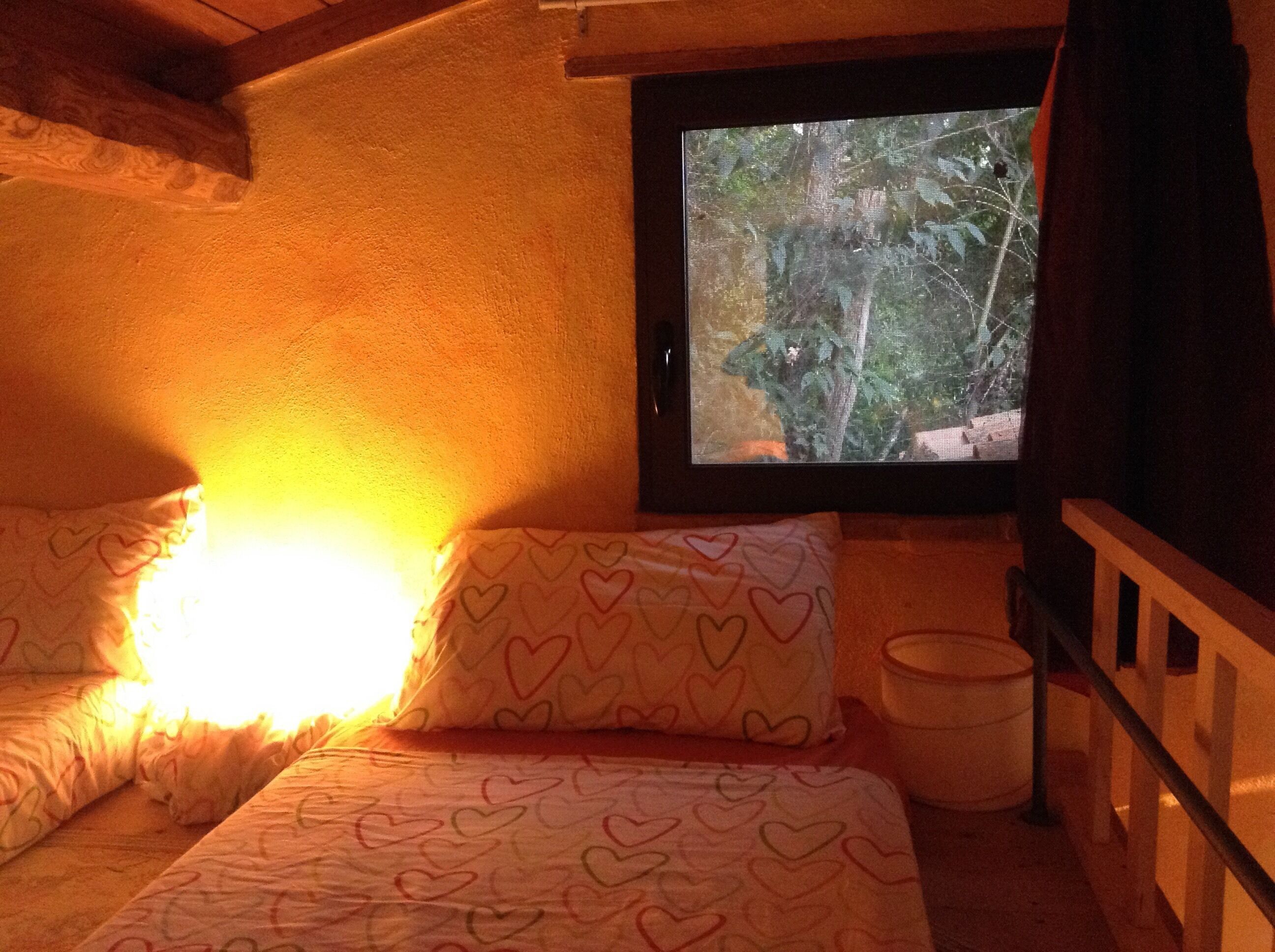 A cozy room with a bed, pillows with heart patterns, a window with a view of trees, and a bucket on the floor.