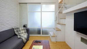 Uma sala de estar moderna com um sofá cinzento, uma mesa de centro de madeira com um aviso de boas-vindas, uma televisão e uma escada com guarda-corpo de vidro.