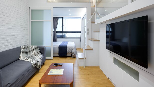 Uma sala de estar moderna com sofá, mesa de centro e televisão de tela plana. Há uma cama com uma janela ao fundo e uma escada que leva a um andar superior.