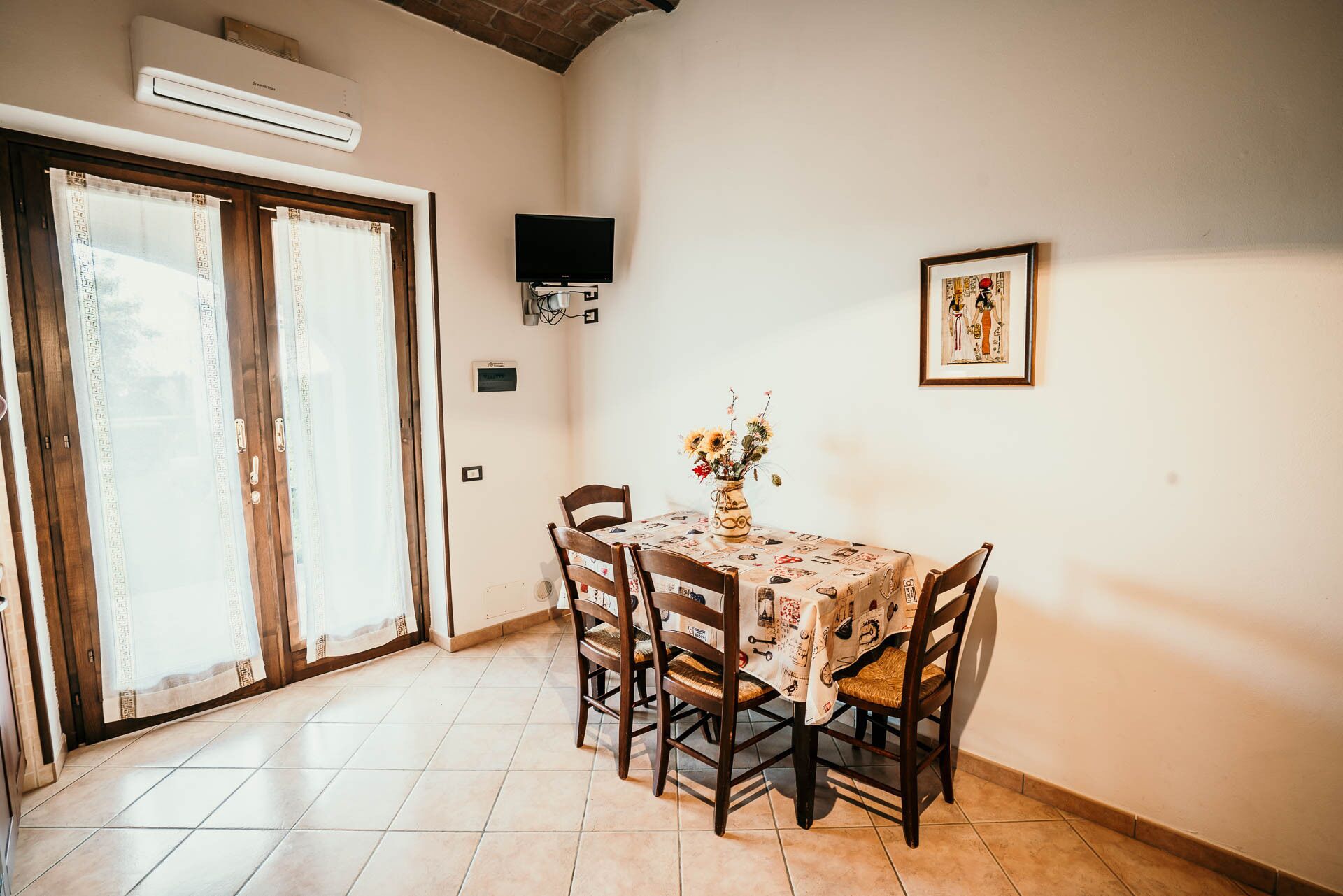 A dining area with a table set for four, surrounded by wooden chairs. There is a vase with flowers on the table, a framed picture on the wall, and an air conditioning unit above the door.