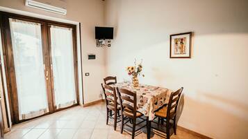 A dining area with a table set for four, surrounded by wooden chairs. There is a vase with flowers on the table, a framed picture on the wall, and an air conditioning unit above the door.