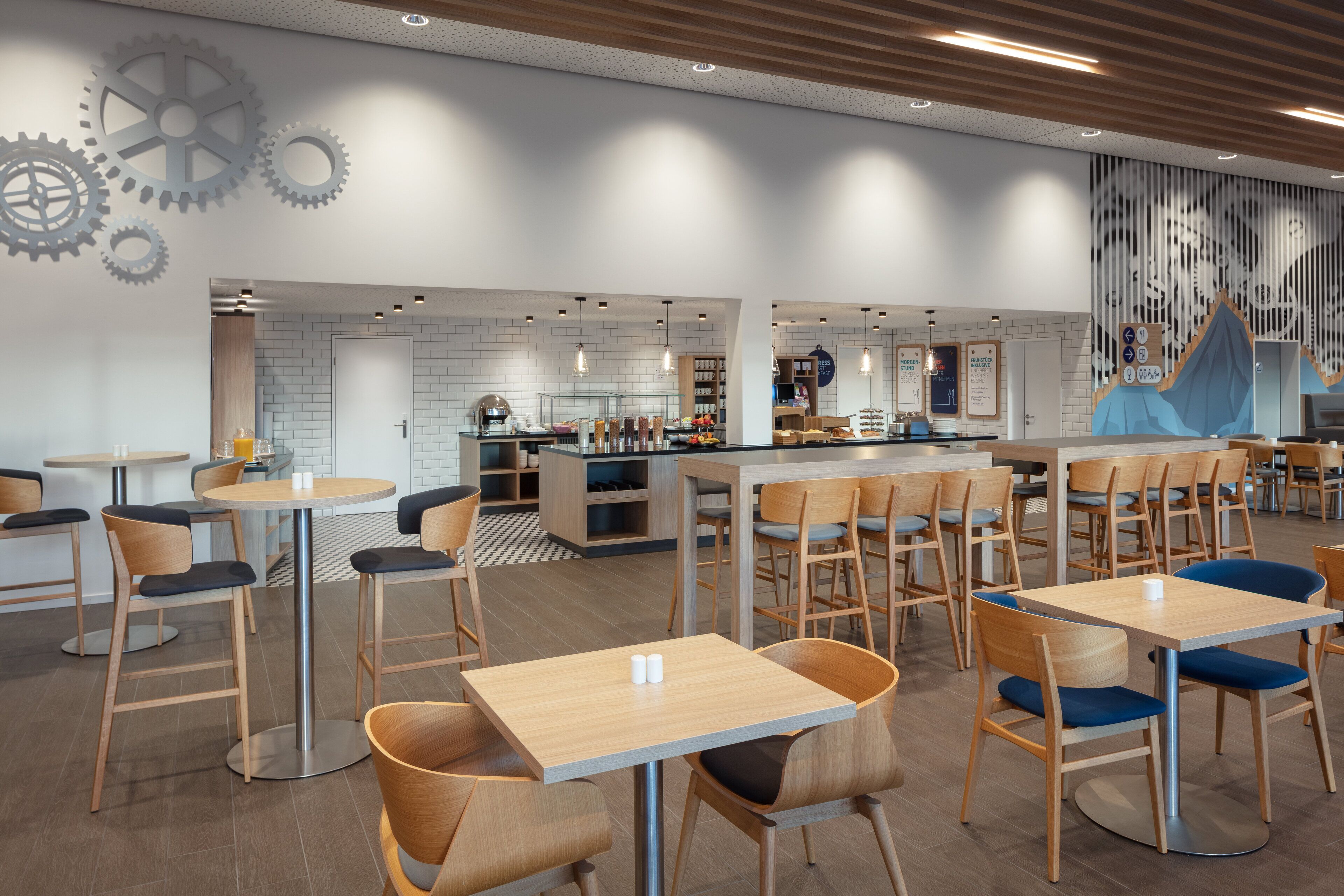 A modern restaurant with wooden tables and chairs, a bar area, and a wall with large gears.