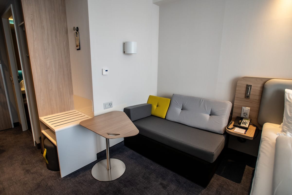 A compact hotel room with a grey sofa, a small wooden table, a bedside table with a phone, and a wall-mounted light.