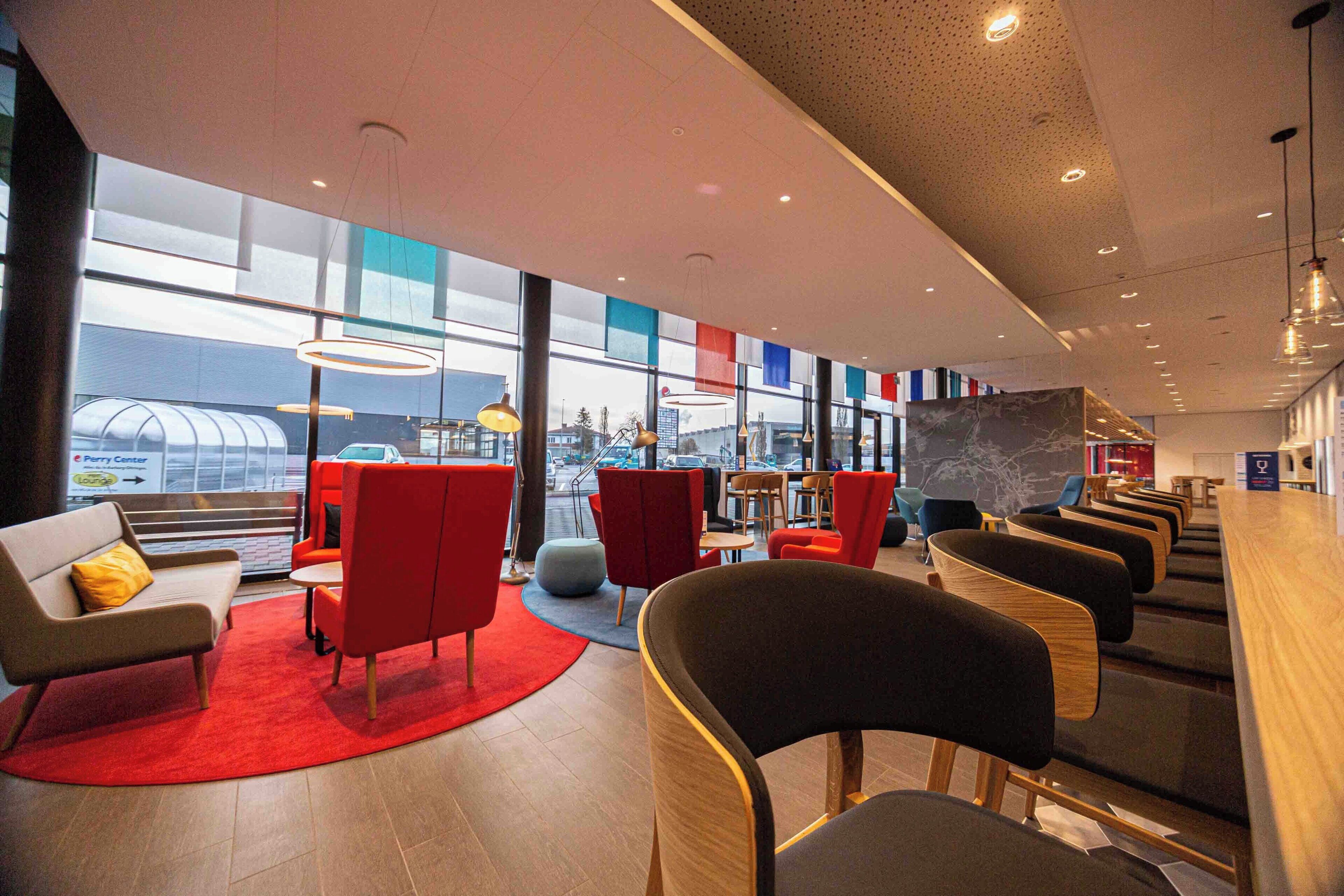 Un área de estar moderna con asientos rojos y grises, ventanas amplias y vistas panorámicas de la ciudad.