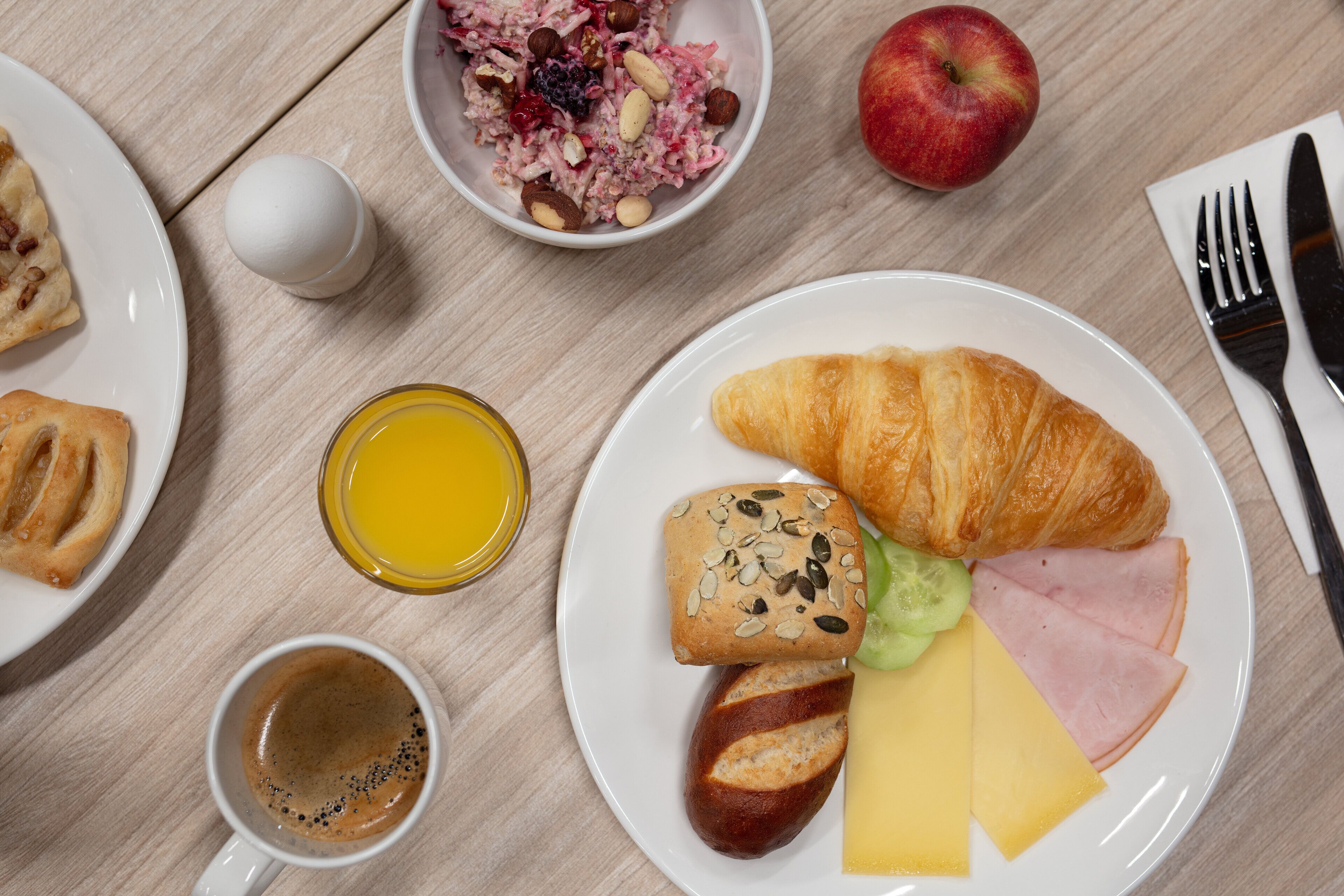 Une table en bois dressée pour le petit-déjeuner, avec des croissants, du fromage, du jambon, du pain, un saladier de fruits, une pomme, un verre de jus d’orange et une tasse de café.