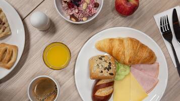 Un desayuno con cruasanes, queso, jamón, pan, un bol de fruta, una manzana, un vaso de jugo de naranja y una taza de café sobre una mesa de madera.