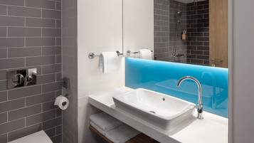 Une salle de bain moderne avec des murs recouverts de carreaux gris, un lavabo blanc agrémenté d’une touche de bleu, des toilettes et un miroir qui reflète l’espace de la douche.