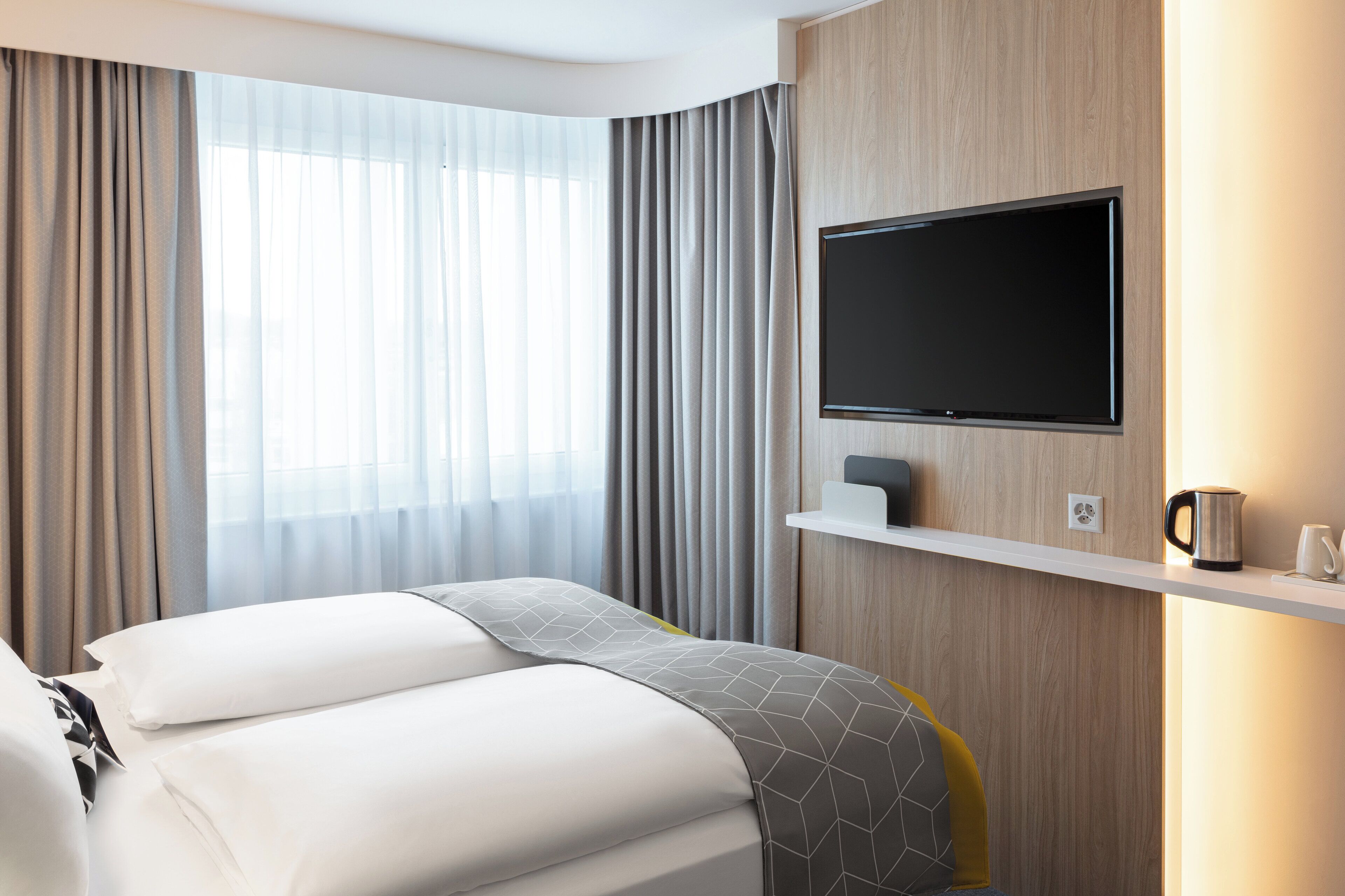 Ein Hotelzimmer mit einem Bett, einem Wand-Fernseher, einer Ablage mit Kaffeemaschine und Tasse sowie Vorhängen.
