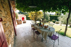 An outdoor patio with a round table and chairs, a stone wall, and a view of a garden and building in the background. - Vacation rental house with pool LIOUX LS2-92 (Lioux)