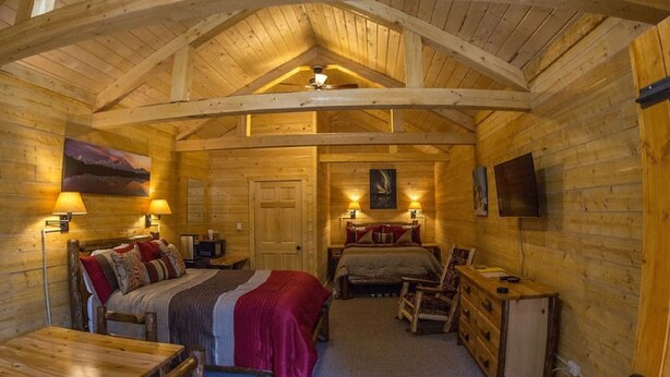 A cozy log cabin bedroom with a large bed, a smaller bed, a desk, and a chair. There is a ceiling fan, a TV mounted on the wall, and a door. - Cozy Bear Log Cabin Tri-Valley  (Healy)