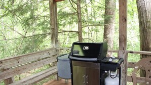 A portable grill with a digital display, a propane tank, and a trash can on a wooden deck surrounded by trees. - Mountaineer Relaxing 2 Bedroom Cabin  (Hico)
