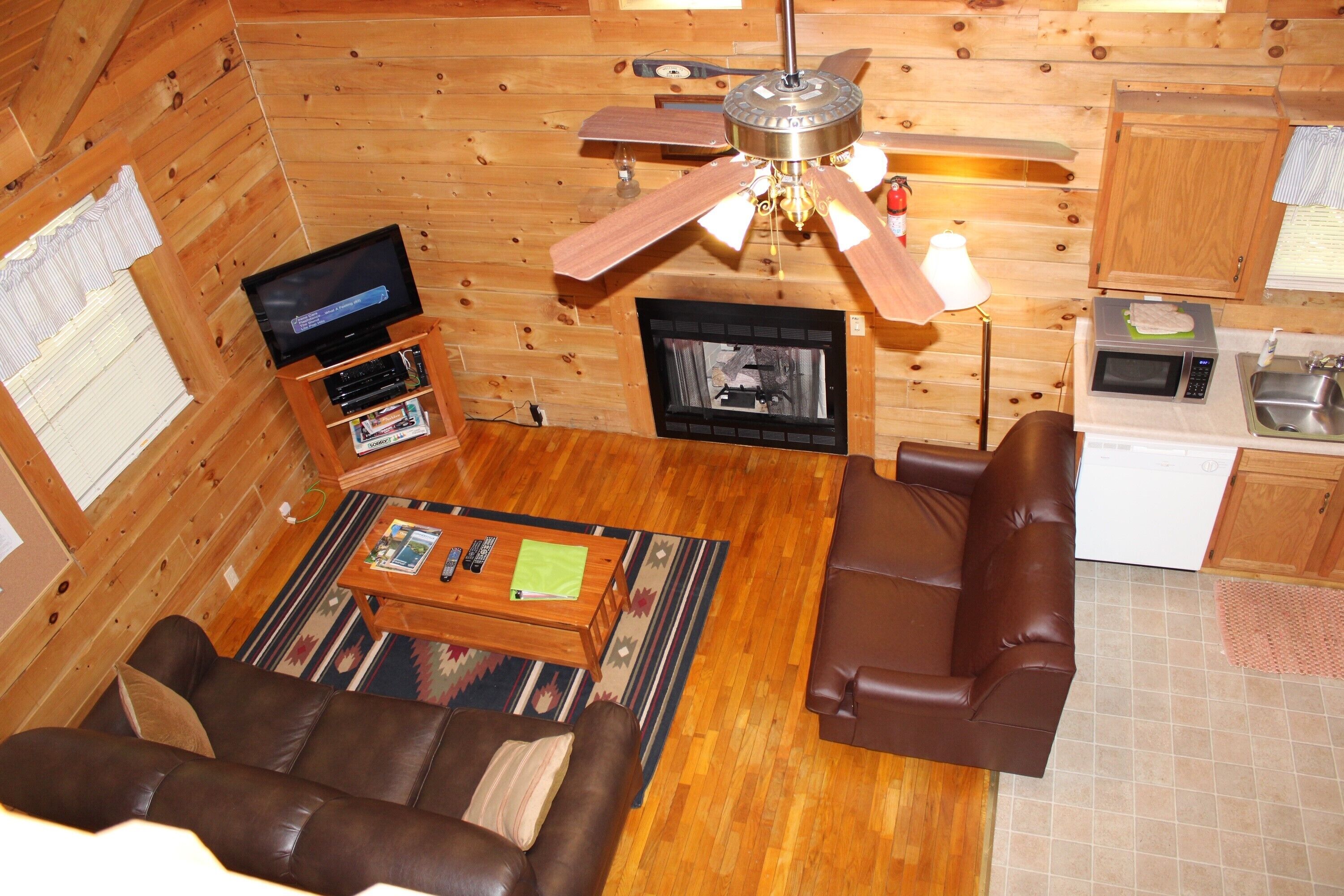 A wooden cabin interior with a living room, sofa, television, fireplace, kitchen area, and ceiling fan.