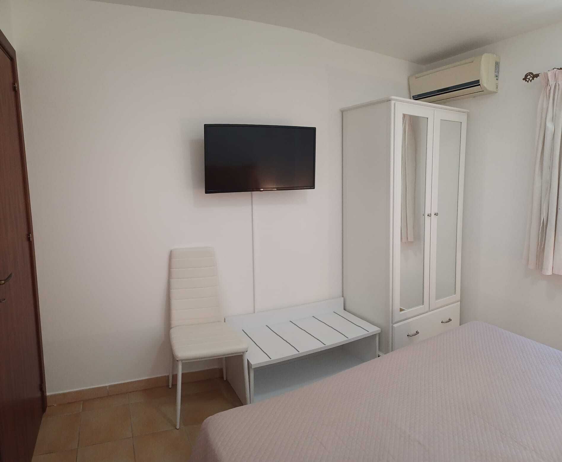 A bedroom with a bed, a white wardrobe, a TV mounted on the wall, and a chair.