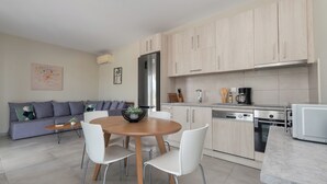 A modern kitchen with light wood cabinets, a stainless steel refrigerator, a dishwasher, and a microwave. There is a dining table with white chairs and a small plant on it.