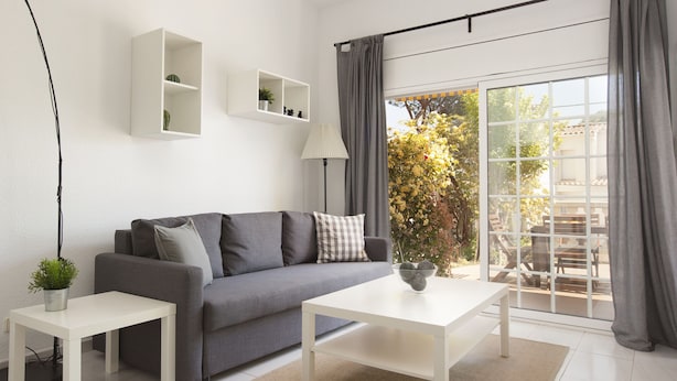 A modern living room with a grey sofa, white coffee table, and side tables. There are white shelves with plants and a lamp.