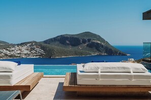 A poolside area with two wooden loungers, one with a mattress and pillows, and a clear view of a mountainous coastline and the sea. - Villa Buse, wonderful sea view. (Kalkan)