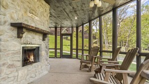 Una chimenea de piedra con fuego, sillas de madera y ventanas amplias con vistas a un paisaje verde.
