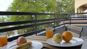 Un balcon avec une table dressée pour le petit-déjeuner, où l’on trouve des assiettes avec une banane, une pomme et une orange, ainsi que des verres de jus d’orange.