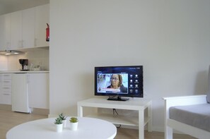 Ein modernes Wohnzimmer mit Flachbildfernseher, einem weißen Couchtisch mit Pflanzen, einem weißen Sofa und einer Küchenzeile im Hintergrund.