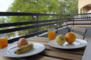 Een balkon met een ontbijttafel, compleet met borden met een banaan, een appel en een sinaasappel, en glazen sinaasappelsap.
